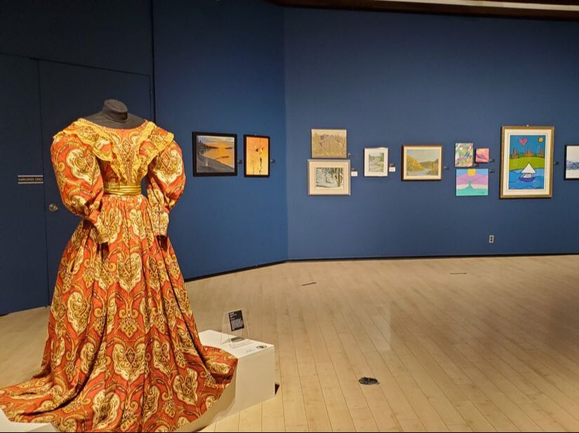 This is a photo of gallery 1. Several paintings are hung up on a blue wall in the background. In the foreground, a historic dress design is displayed on a mannequin.
